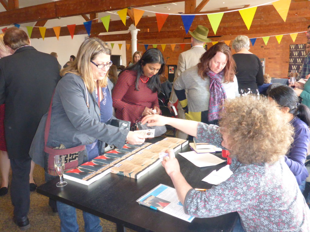 3 people standing around book stall, one paying for a book and the others browsing. People and bunting in background.