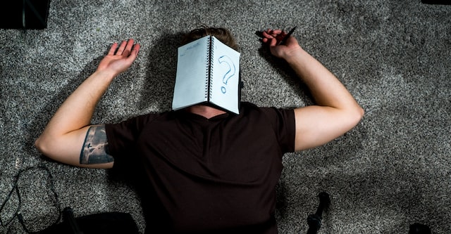 Man lying on floor with writing book over his face and pen in hand,. The book has a question mark on the front cover